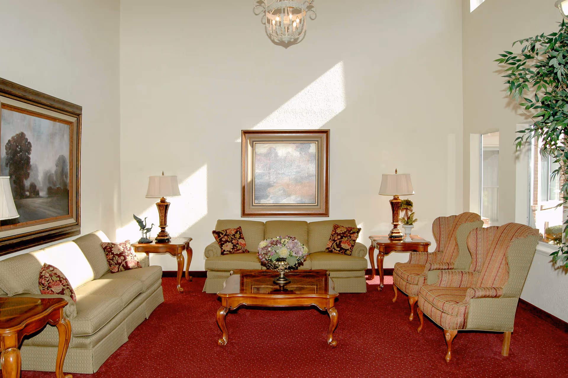 A bright living room with two beige sofas and two patterned armchairs arranged around a wooden coffee table with a floral centerpiece. The room has red carpet, two side tables with lamps, framed artwork on the walls, and a large window letting in natural light.