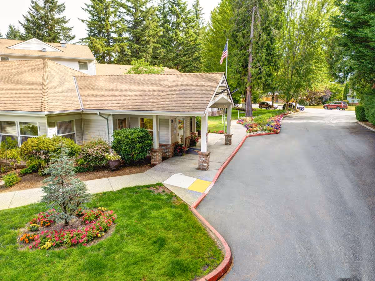 Exterior view of a senior living facility entrance with a covered porch supported by brick columns, surrounded by well-maintained landscaping including green grass, colorful flowers, shrubs, and tall trees. A paved driveway curves around the building with parked cars visible in the background.