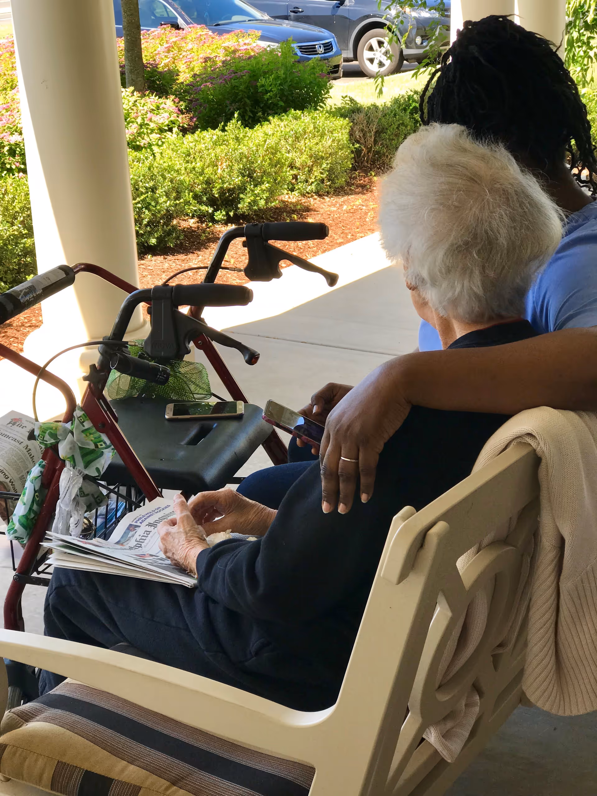 An elderly person with white hair sitting on a bench outdoors, holding a newspaper, with a walker nearby. A younger person with dark braided hair sits beside them, with an arm around the elderly person, both looking at a smartphone. There are bushes and parked cars in the background.