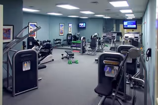 A fitness room equipped with various exercise machines including weight machines, treadmills, and stationary bikes. The room has light blue walls, a gray carpeted floor, and mirrors on one wall. Two televisions are mounted on the walls, and there are small green dumbbells on the floor.