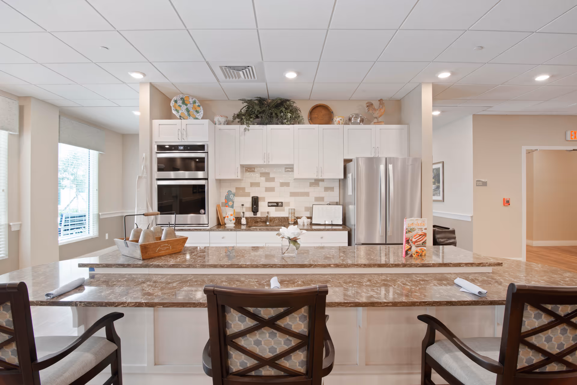 A bright and modern kitchen area with white cabinets, stainless steel double oven and refrigerator, and a marble countertop island with three cushioned chairs. Decorative items and plants are placed on top of the cabinets. The room has large windows letting in natural light and recessed ceiling lights.
