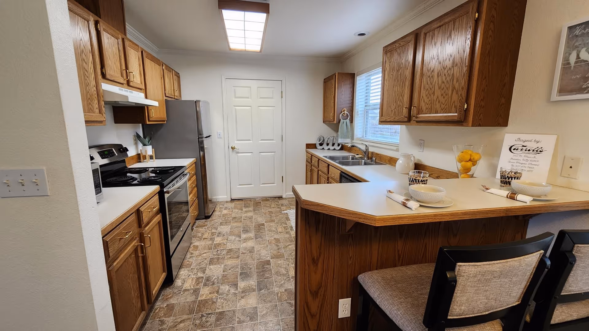 A well-lit kitchen with wooden cabinets, a stainless steel refrigerator, stove, and microwave. There is a white countertop with two place settings, each with a bowl, cup, and napkin. A window above the sink lets in natural light, and the floor has a patterned tile design.