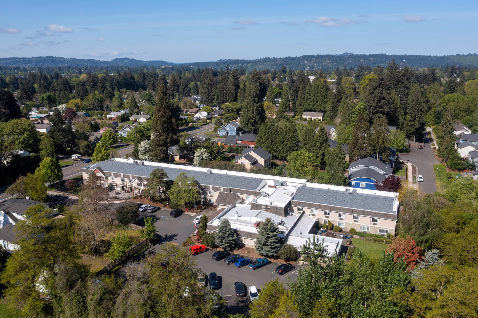Aerial view of Royalton Place senior living facility surrounded by trees and residential houses, with a parking lot in front and a forested area in the background under a partly cloudy sky.