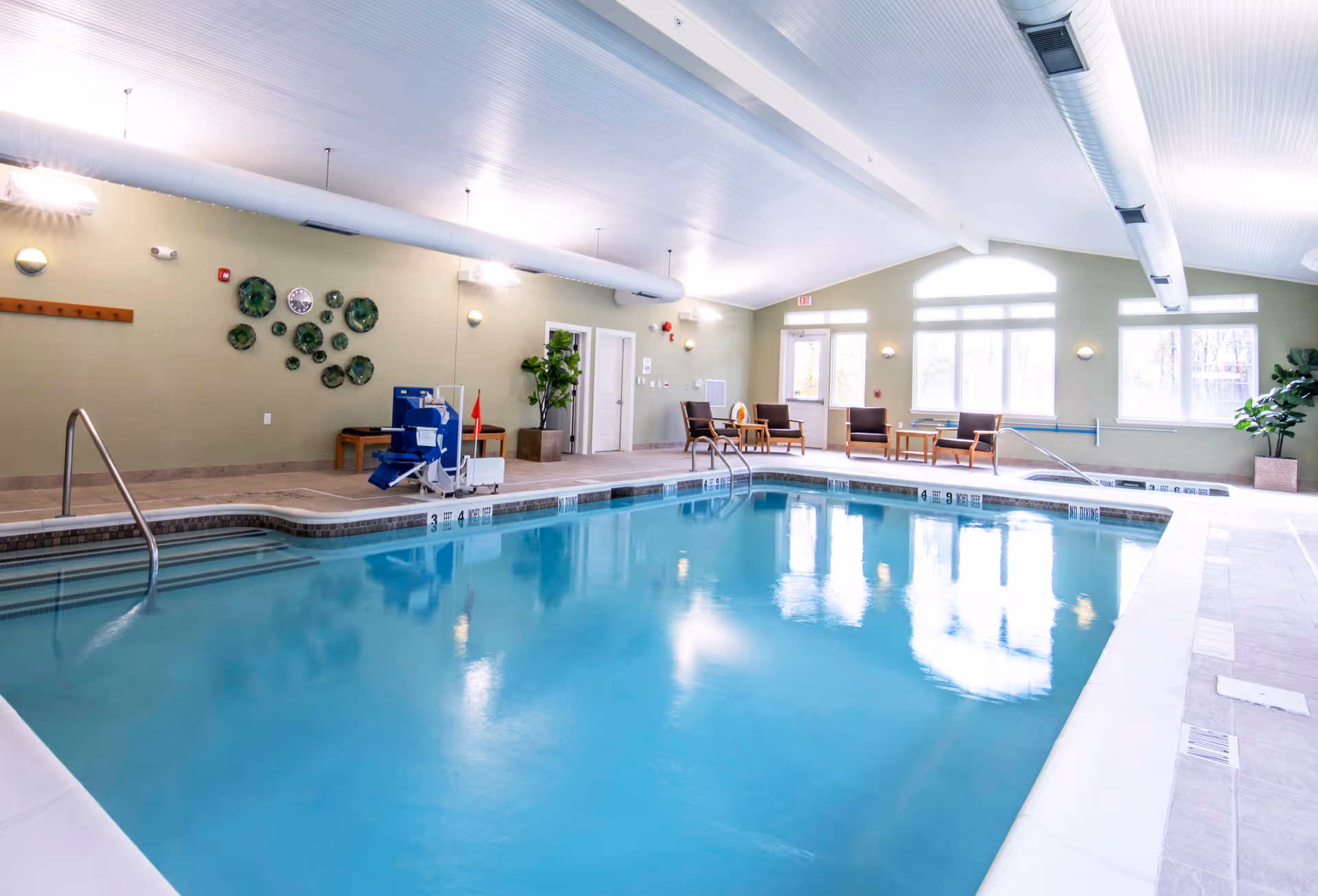 Bright indoor swimming pool with seating, a pool lift, and large windows in a recreation room.