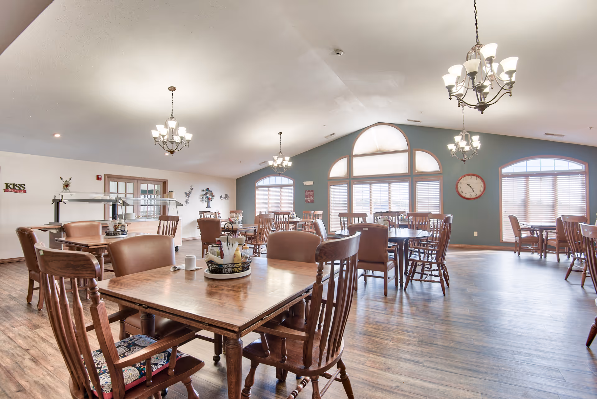 A spacious dining room with multiple wooden tables and chairs arranged neatly. The room has large arched windows with blinds, allowing natural light to fill the space. Several chandeliers hang from the ceiling, providing additional lighting. The walls are painted in a combination of light and muted blue tones, and there is a red clock on one wall. The floor is covered with wood-like flooring, and there are decorative items on the walls and tables.