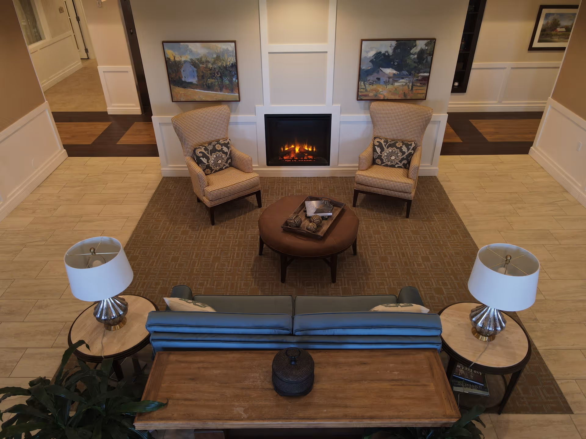 A cozy senior living facility common area with two patterned armchairs and a round ottoman in front of a lit fireplace. There is a blue sofa with two side tables holding lamps, and framed landscape paintings on the wall above the fireplace.