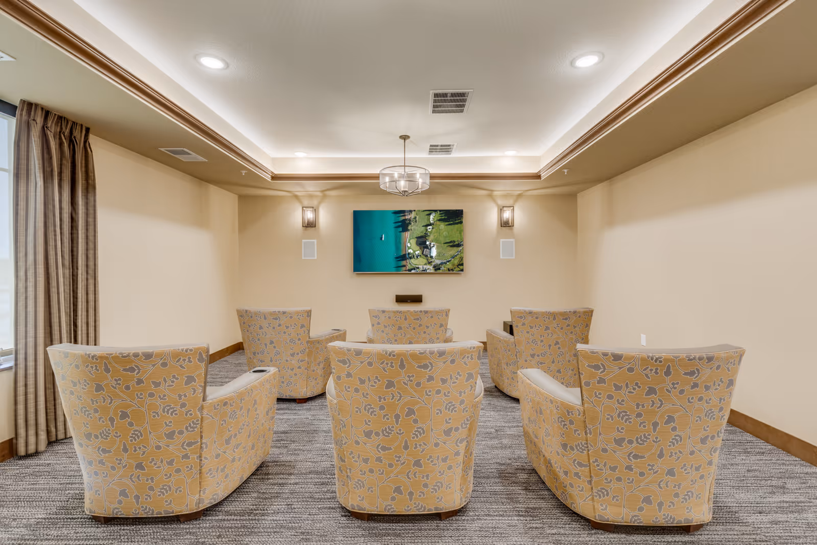 A small media room with six patterned armchairs arranged in two rows facing a wall-mounted flat-screen TV. The room has beige walls, a carpeted floor, recessed ceiling lights, and a window with brown curtains on the left side.