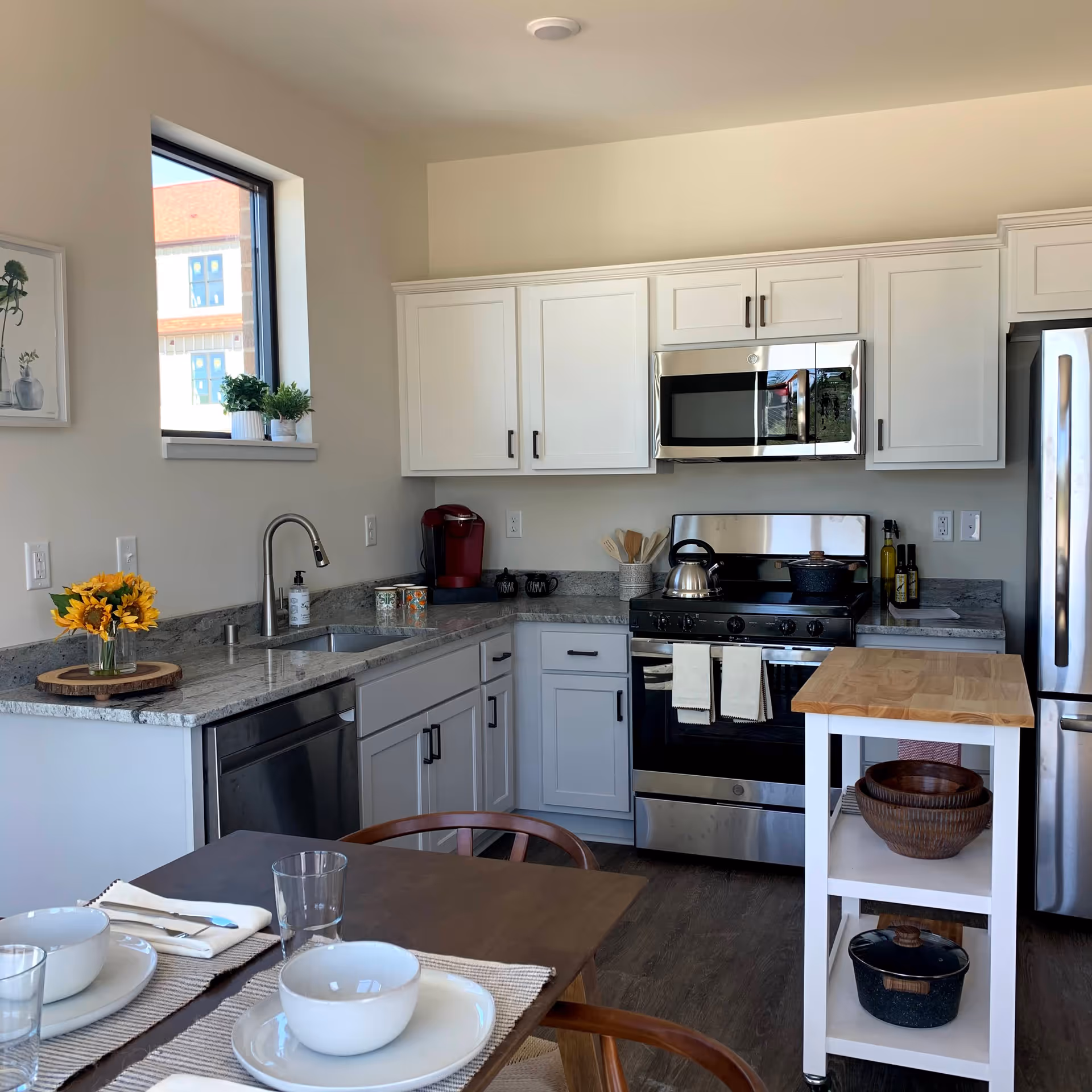 A modern kitchen with white cabinets, stainless steel appliances including a microwave, stove, dishwasher, and refrigerator. There is a small wooden island with shelves holding bowls and a pot. A dining table with place settings including bowls, plates, glasses, and napkins is in the foreground. A window with two small potted plants and a framed picture on the wall are visible.