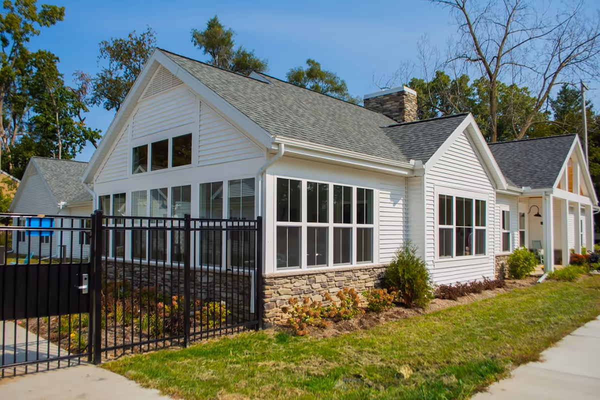 White single-story building with large windows, stone foundation and a black metal fence in a landscaped yard.