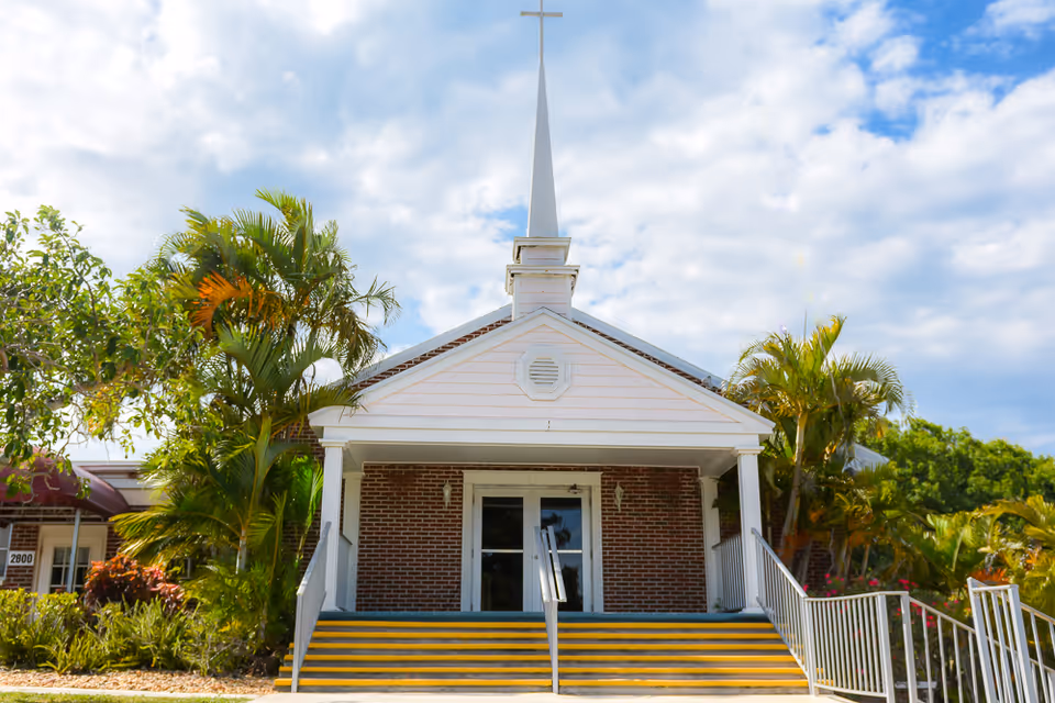 Front exterior view of a brick building with white columns and a tall white steeple topped with a cross, surrounded by palm trees and greenery under a partly cloudy sky.