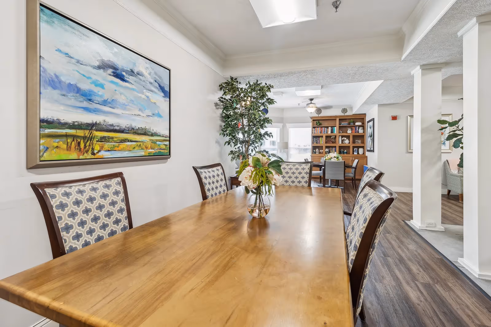 Long wooden dining table with patterned chairs, a vase of flowers, wall art, and a communal seating area and bookshelf in a bright dining room.