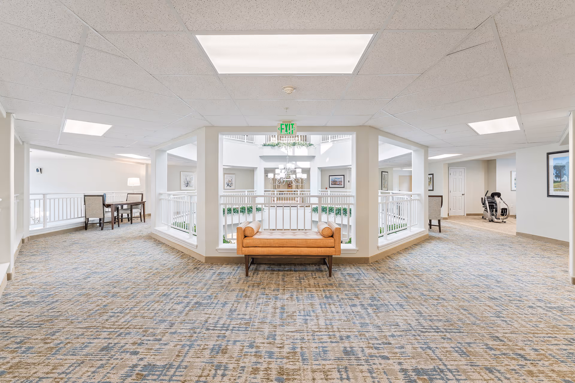Bright and spacious interior hallway area in a senior living facility with patterned carpet flooring, a tan cushioned bench in the center, white railings overlooking an atrium, and seating areas with chairs and tables on either side. The ceiling has recessed lighting panels and there are framed pictures on the walls.