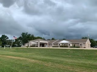 Single-story brick building with a covered entrance, flagpole, and wide grassy lawn under a cloudy sky.