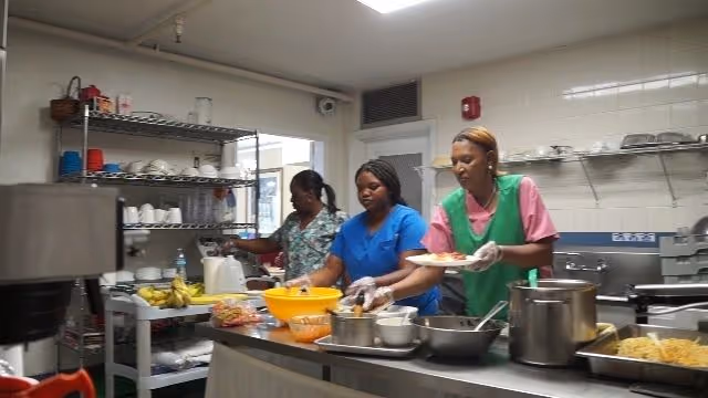 Three staff members preparing and plating food in a commercial kitchen with pots, bowls, and shelving.