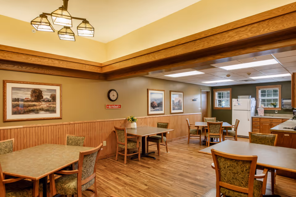 Dining room with several tables and upholstered chairs, wood-paneled walls, framed artwork, and a small kitchenette with a refrigerator.