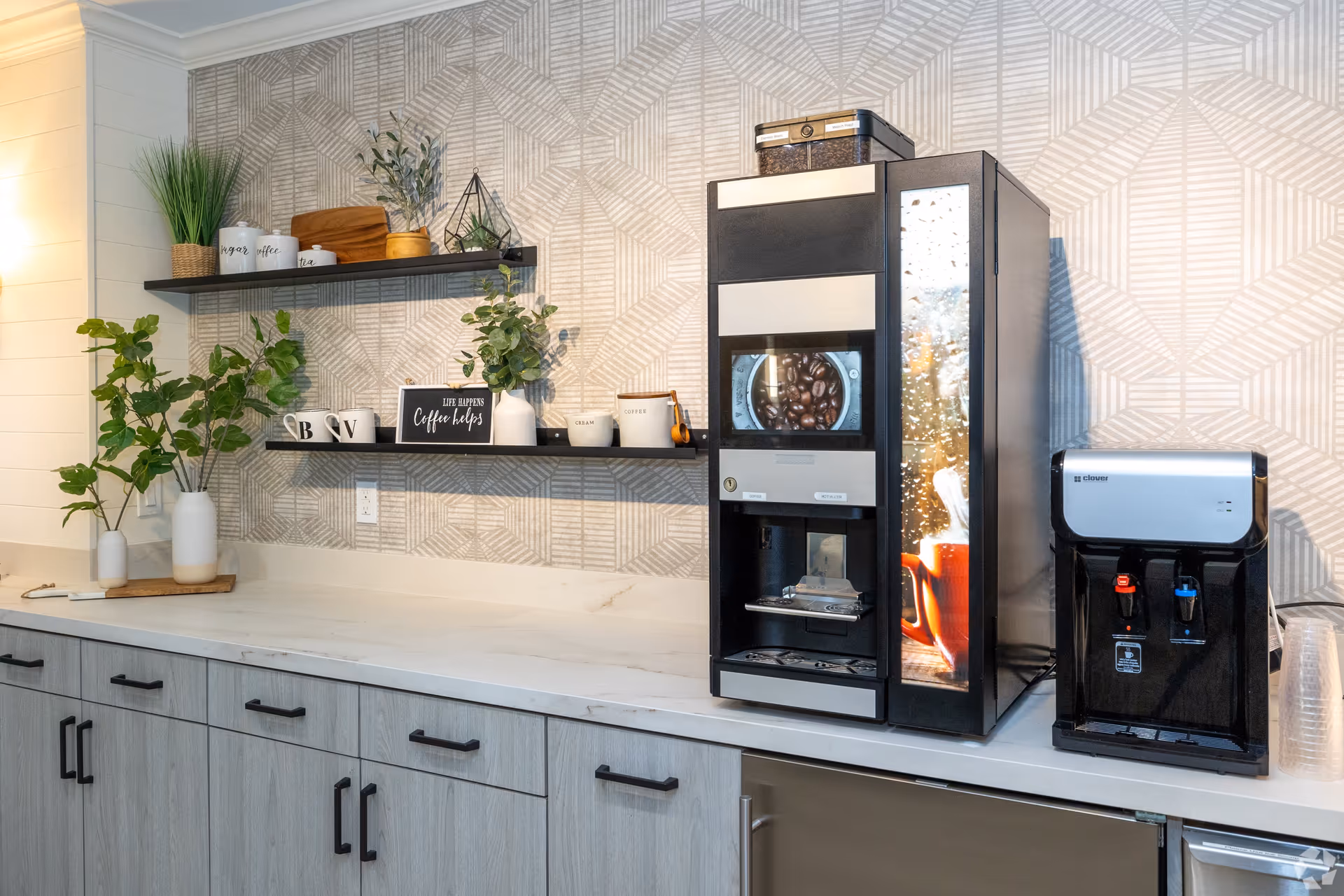 A modern coffee station with a coffee machine and water dispenser on a white countertop. Above the counter are two black floating shelves holding decorative plants, mugs, and containers. The wall behind has a geometric patterned wallpaper.