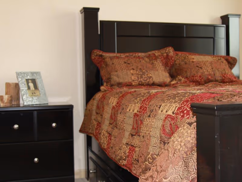 A bedroom with a dark wood bed topped by a patterned red and gold bedspread and pillows, next to a black nightstand holding a framed photo.