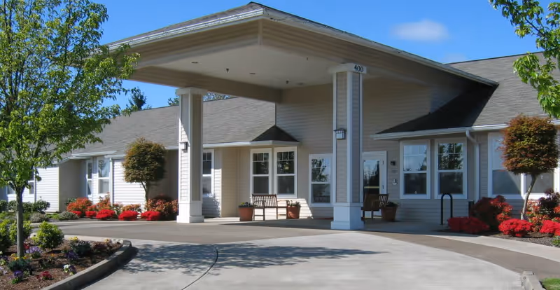 Exterior view of a single-story assisted living facility building with a covered entrance supported by two columns. The building is surrounded by landscaped gardens with small trees, shrubs, and red flowers. There are benches and potted plants near the entrance under a clear blue sky.