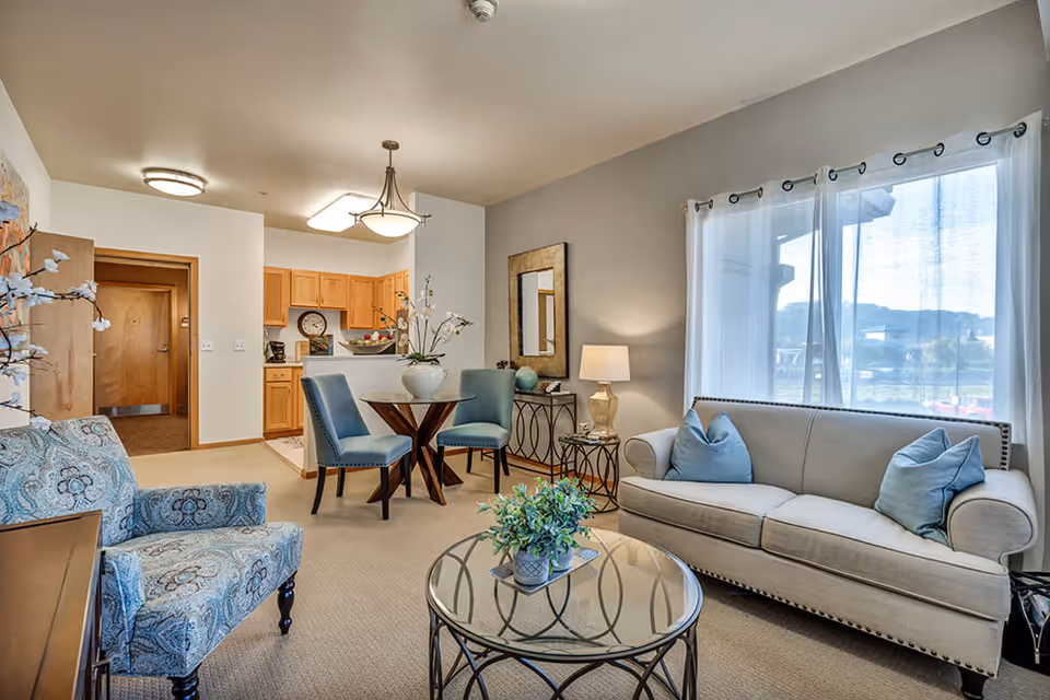 A cozy living room and dining area in a senior living facility. The room features a beige sofa with blue pillows, a patterned armchair, a round glass coffee table with a small plant, and a small dining table with two blue chairs. The walls are painted light gray, and there is a large window with sheer white curtains letting in natural light. The kitchen with wooden cabinets is visible in the background.