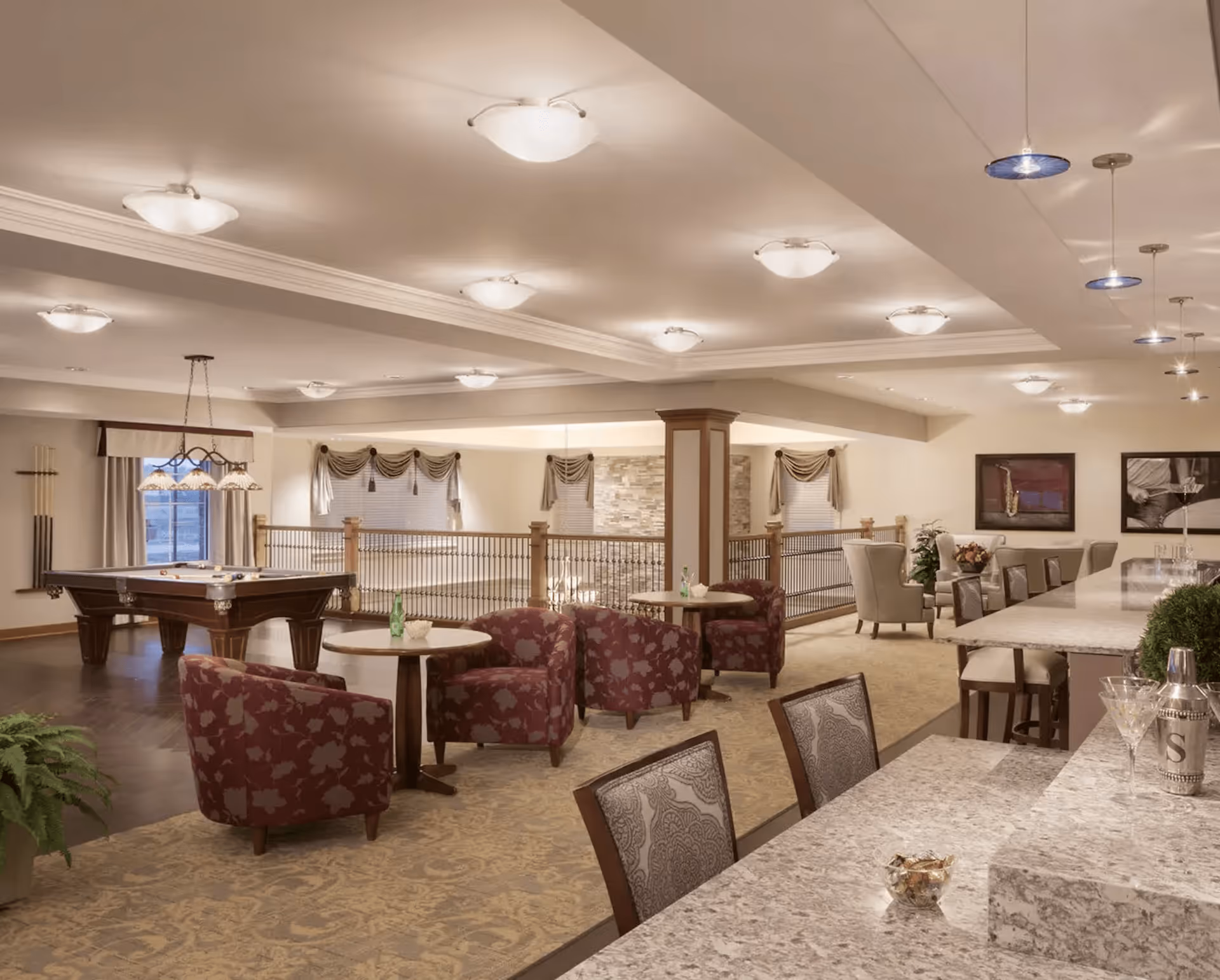 A spacious and well-lit common area in a senior living facility featuring a pool table, several seating areas with patterned armchairs and small round tables, a long granite countertop with bar stools, and decorative lighting fixtures on the ceiling. The room has beige walls, carpeted and wooden floors, and framed artwork on the walls.