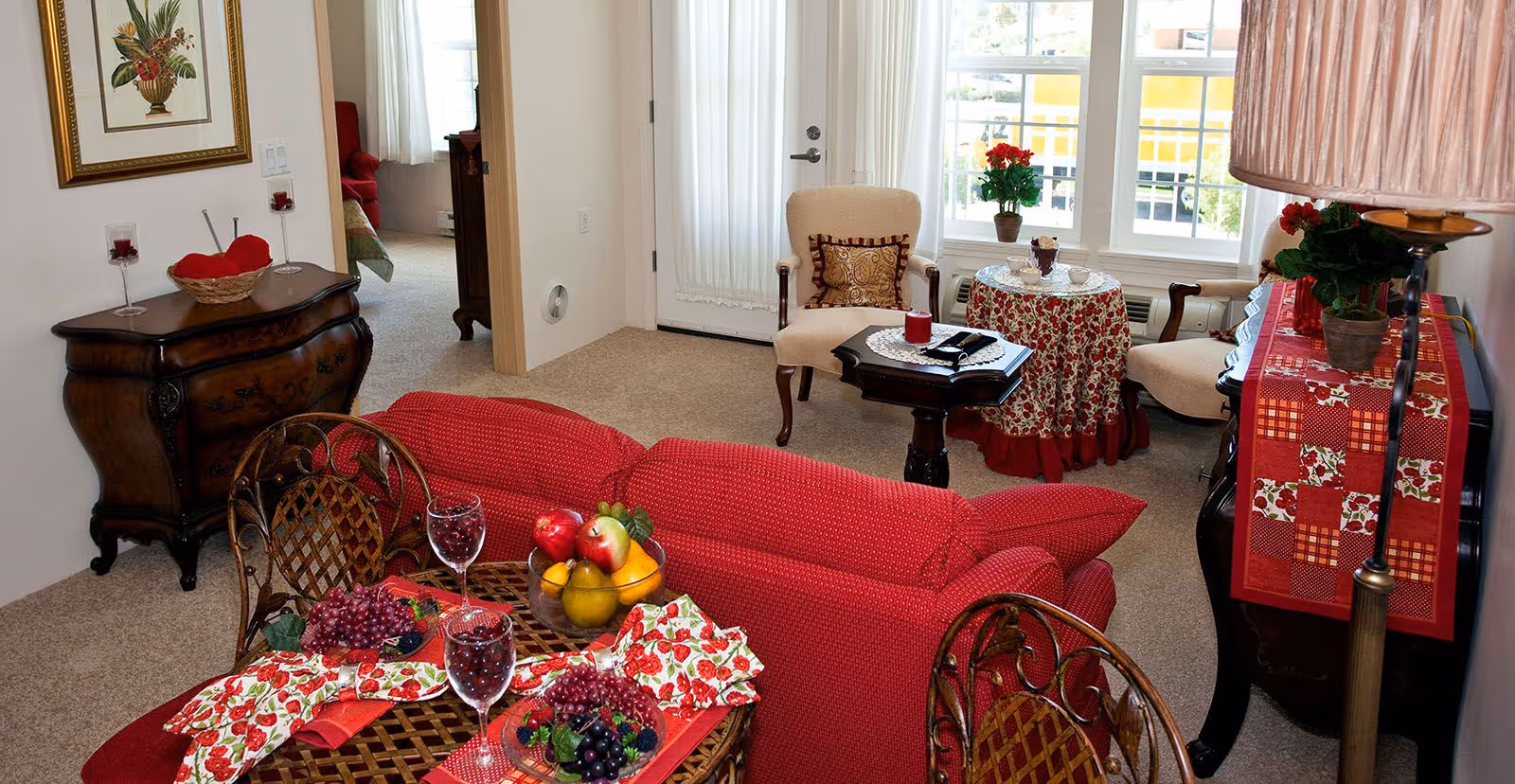 A cozy living room with a red sofa, a small round table covered with a floral tablecloth, two beige armchairs, and a wooden side table. The room has a carpeted floor, a door with a sheer curtain, and windows letting in natural light. There are decorative items including a bowl of fruit, wine glasses, and potted plants on the tables and furniture.