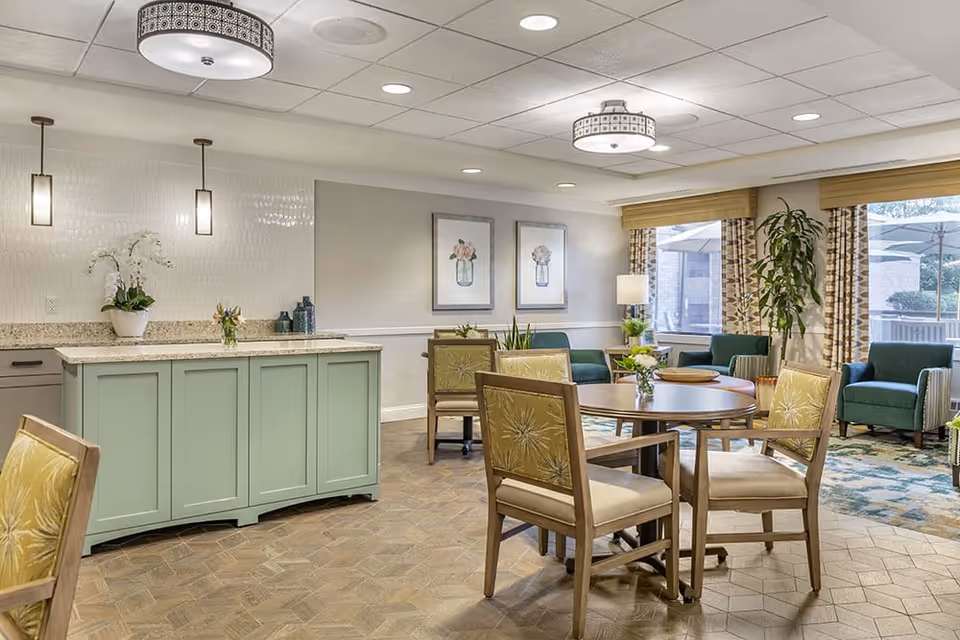 A bright and inviting common area with a round wooden table surrounded by four cushioned chairs with floral upholstery. In the background, there is a green cabinet with a granite countertop, decorated with plants and flowers. The room features large windows with patterned curtains, green armchairs, framed floral artwork on the walls, and ceiling lights providing warm illumination.