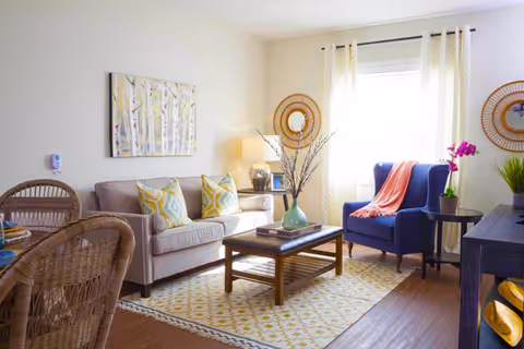 A bright and cozy living room with a beige sofa adorned with patterned yellow and gray pillows, a wooden coffee table with a green vase and books, a blue armchair with a coral throw blanket, a side table with a purple orchid, and two round wicker mirrors on the wall. The room has light-colored walls, a window with sheer curtains, and a patterned yellow and white rug on a wooden floor.