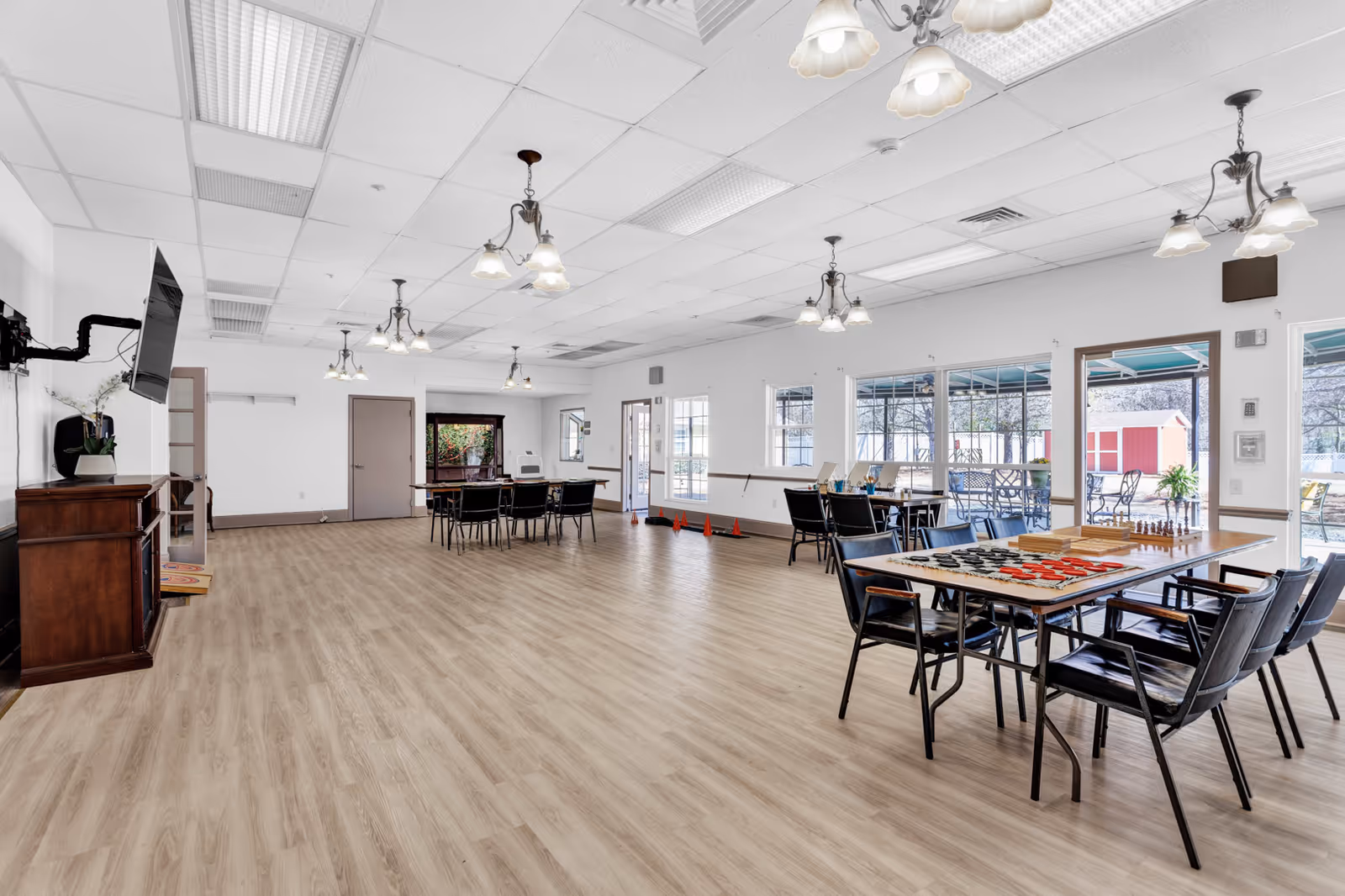 A spacious, well-lit activity room with wooden flooring and multiple tables and chairs arranged for group activities. The room has large windows and glass doors leading to an outdoor patio area. There are board games and checkers set up on one of the tables. The ceiling has multiple light fixtures, and a flat-screen TV is mounted on the wall above a wooden cabinet.