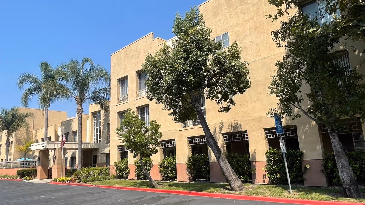 Front exterior of a beige multi-story senior living building with palm trees, an entrance canopy, and landscaped curb.