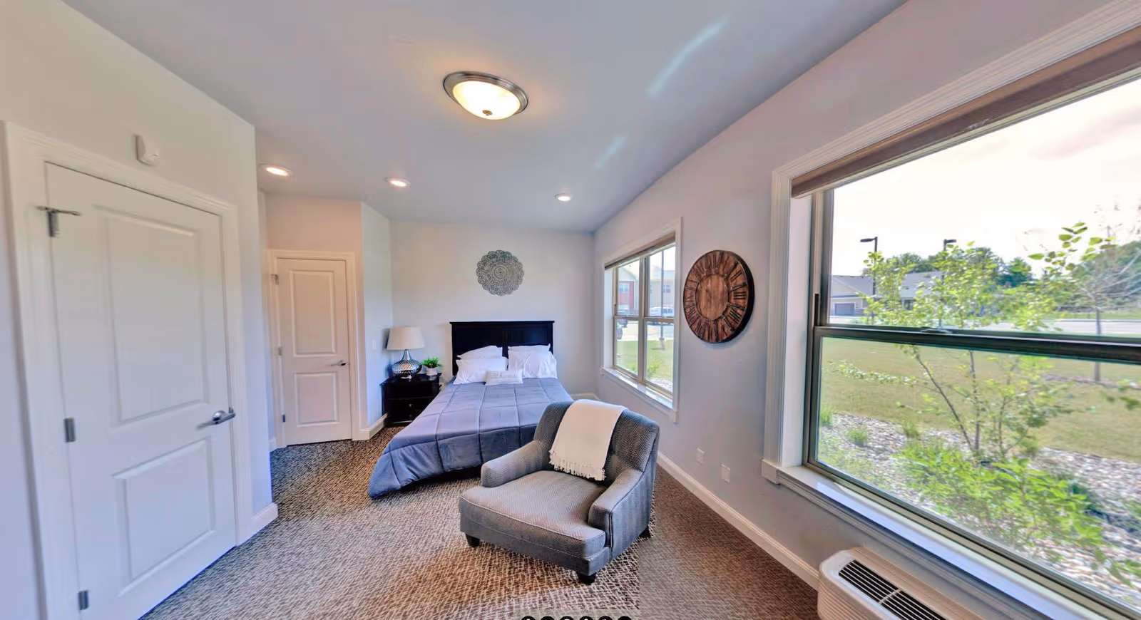 Bright bedroom featuring a bed with a nightstand, an upholstered armchair, and large windows overlooking landscaping.