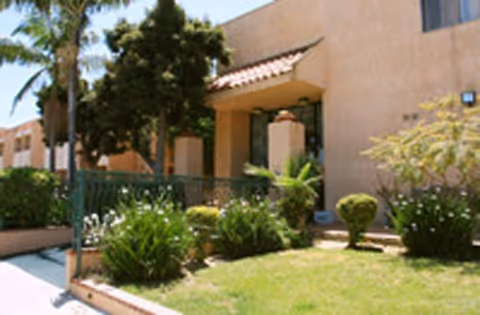 Exterior view of a beige stucco building with a tiled roof overhang above the entrance, surrounded by green shrubs, small trees, and a well-maintained lawn under a clear sky.