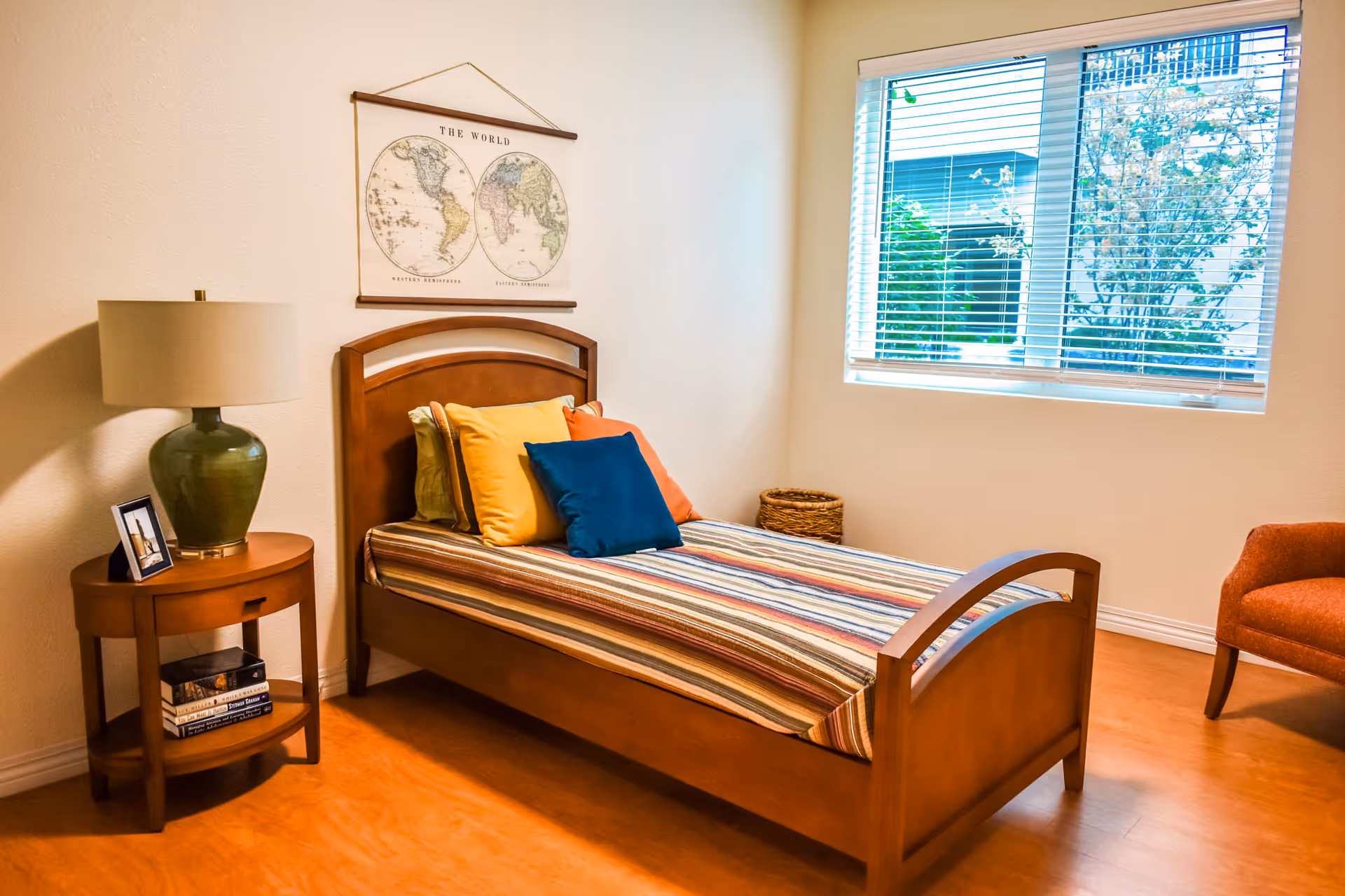 Single wooden bed with striped bedding and colorful pillows in a bright bedroom with a side table, lamp, window, and wall map.