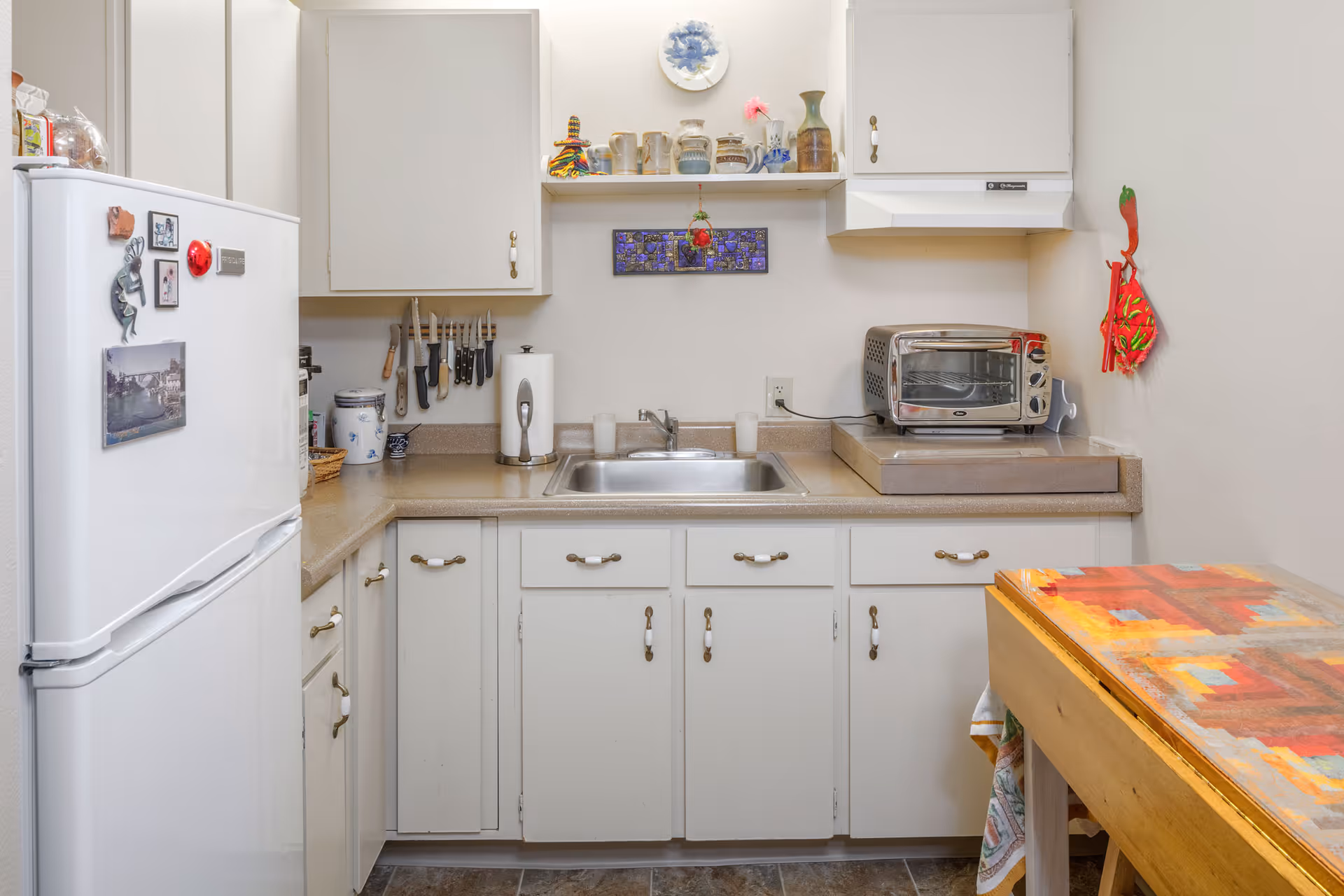 A small kitchen with white cabinets and beige countertops. There is a white refrigerator on the left with magnets and pictures on it. Above the countertop, there are knives hanging on a magnetic strip, a paper towel holder, and various ceramic jars and decorative items on a shelf. A stainless steel sink is centered under the shelf. To the right of the sink, there is a toaster oven on the counter. On the far right, part of a wooden table with a colorful patterned tablecloth is visible. The walls are light-colored, and the floor has dark tiles.