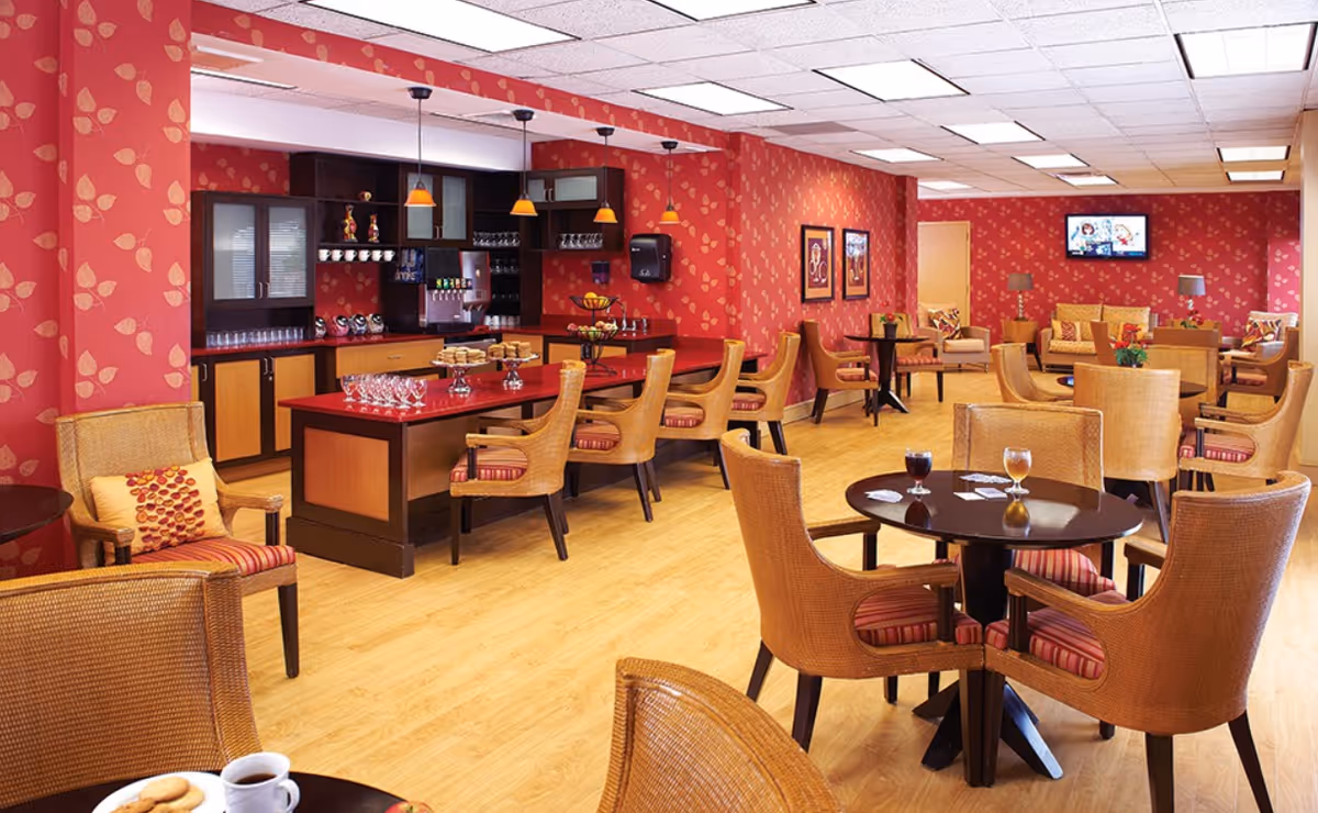 A cozy dining and lounge area with red patterned wallpaper and wooden flooring. The room features multiple round tables with wicker chairs that have red striped cushions. A counter with a red countertop is set with glasses and plates of cookies, with hanging pendant lights above. In the background, there is a seating area with armchairs and a sofa, and a wall-mounted TV.