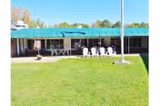 Single-story building with a green roof facing a grassy courtyard with a flagpole and patio seating.
