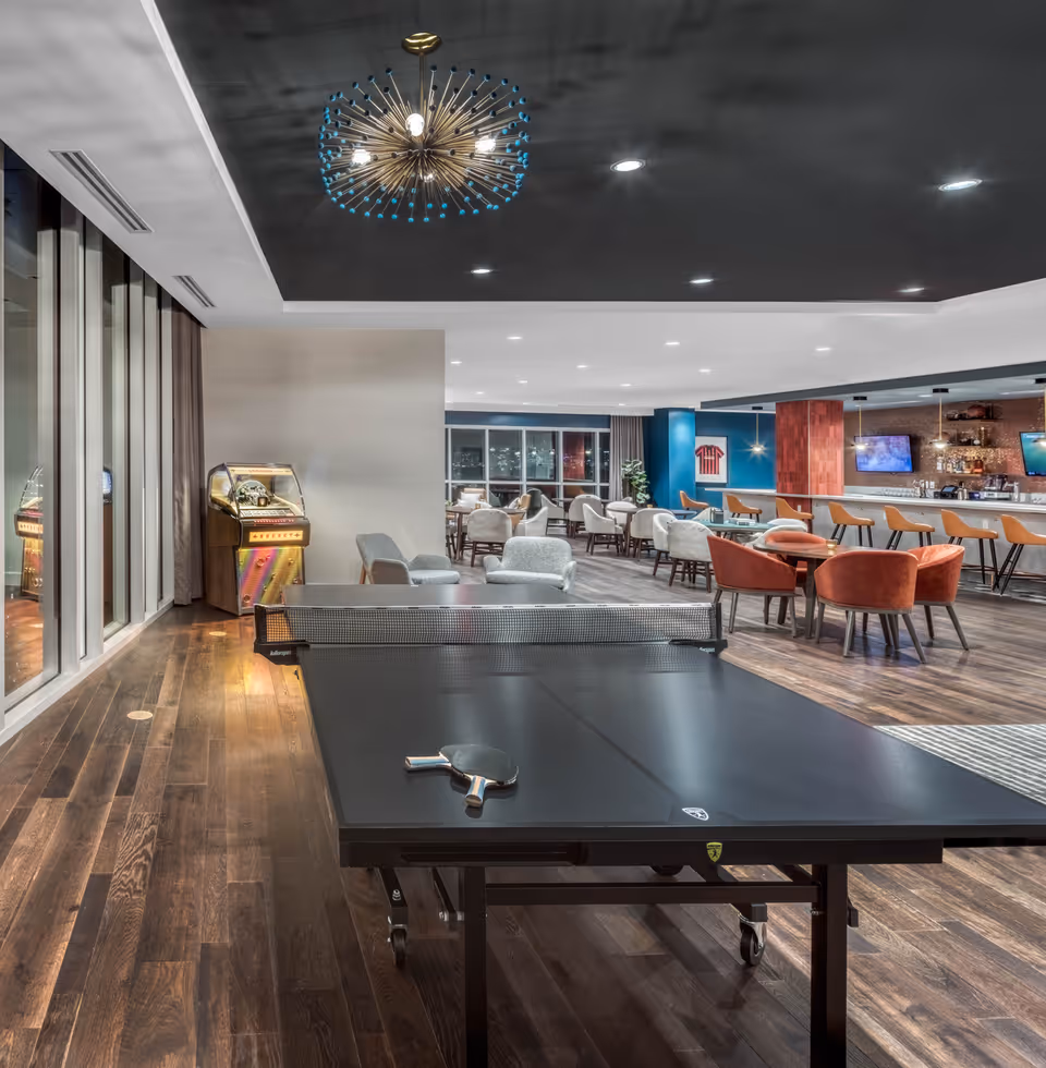 A modern senior living common area featuring a black ping pong table with paddles, a vintage jukebox, multiple seating arrangements including gray and orange chairs, a bar with stools, and large windows with city views. The ceiling is black with recessed lighting and a decorative chandelier.