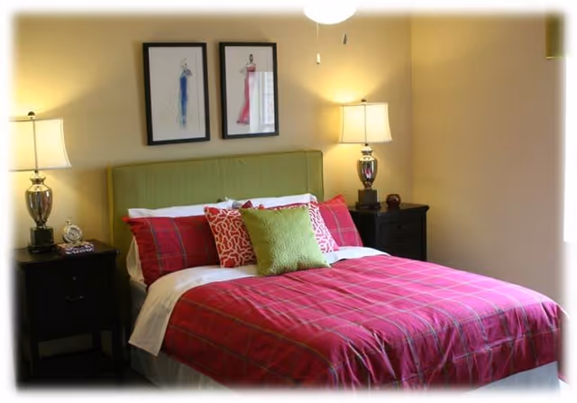 A cozy bedroom with a bed featuring a green upholstered headboard, red plaid bedding, and multiple decorative pillows in red and green. Two black nightstands flank the bed, each with a lamp and small decorative items. Two framed fashion sketches hang on the wall above the bed.