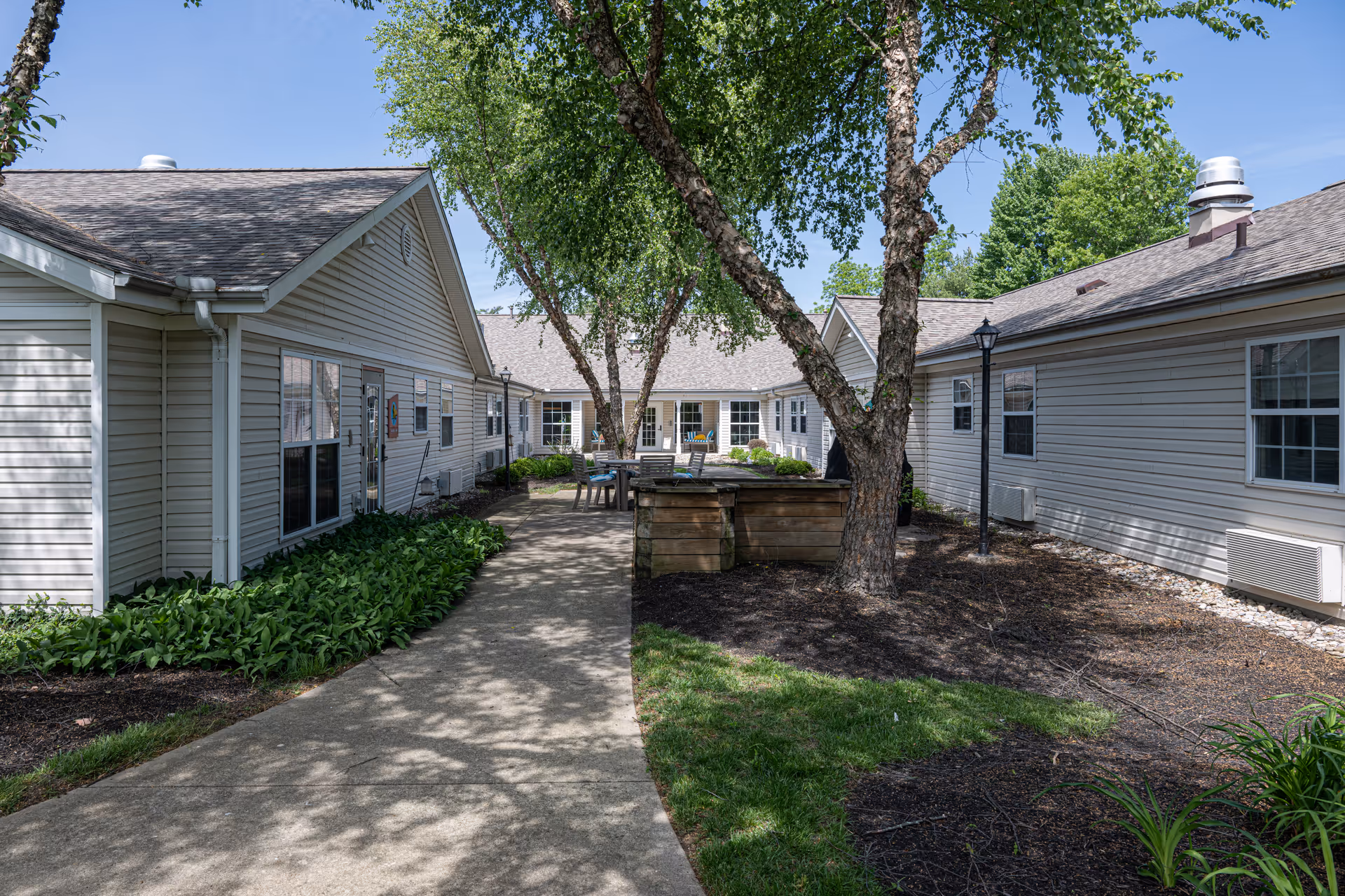 Outdoor courtyard area of a senior living facility with a paved walkway, green plants along the sides, and several trees providing shade. The courtyard is surrounded by single-story buildings with beige siding and multiple windows. There is a wooden planter box and outdoor seating visible in the center of the courtyard under the trees.