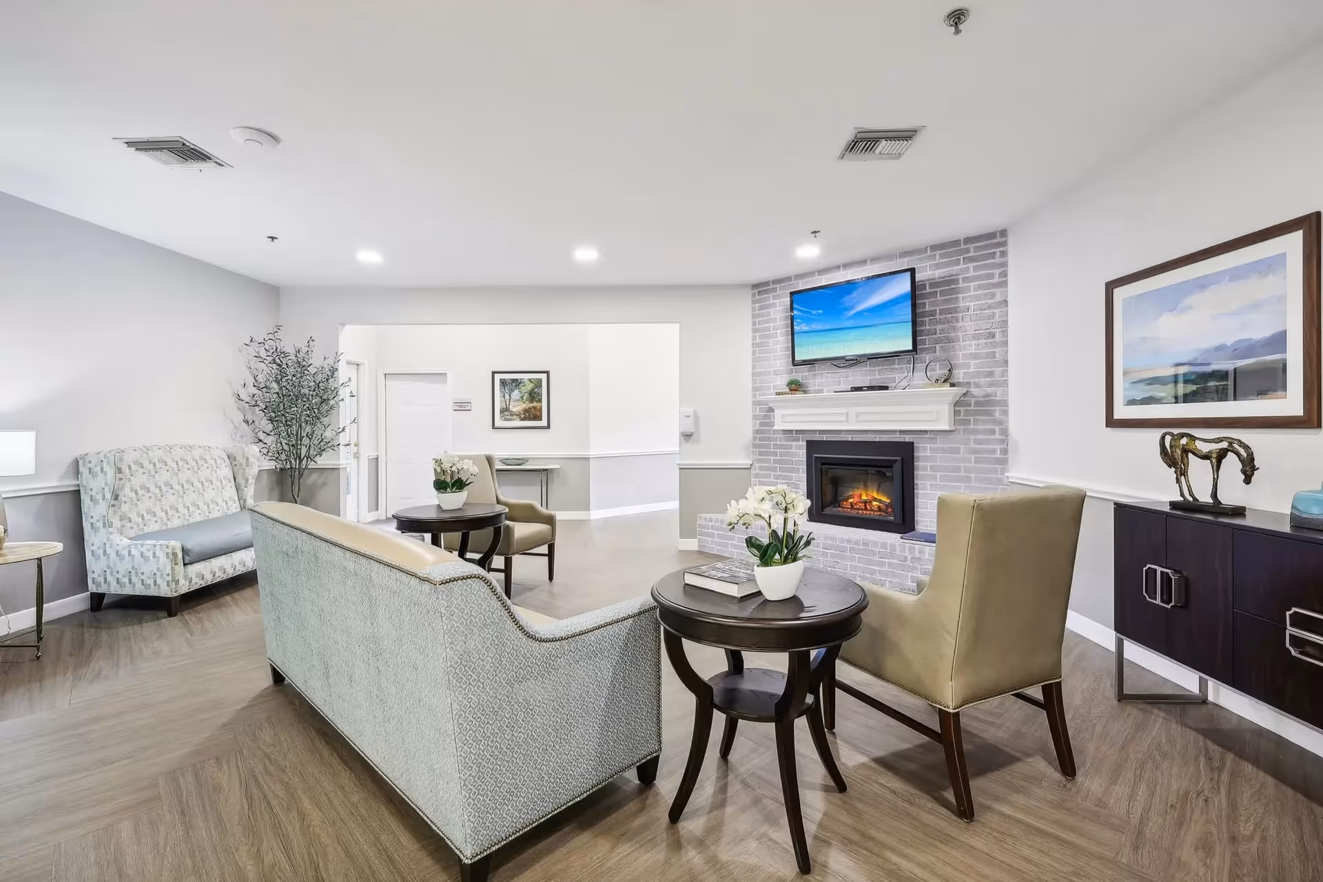 A bright and modern living room area with a light blue patterned sofa, beige armchair, and a small round table with a white flower pot. There is a gray brick fireplace with a flat-screen TV mounted above it displaying a beach scene. The room has light wood flooring, white walls, and decorative artwork on the walls. A dark wooden sideboard with a sculpture and a framed picture is on the right side.