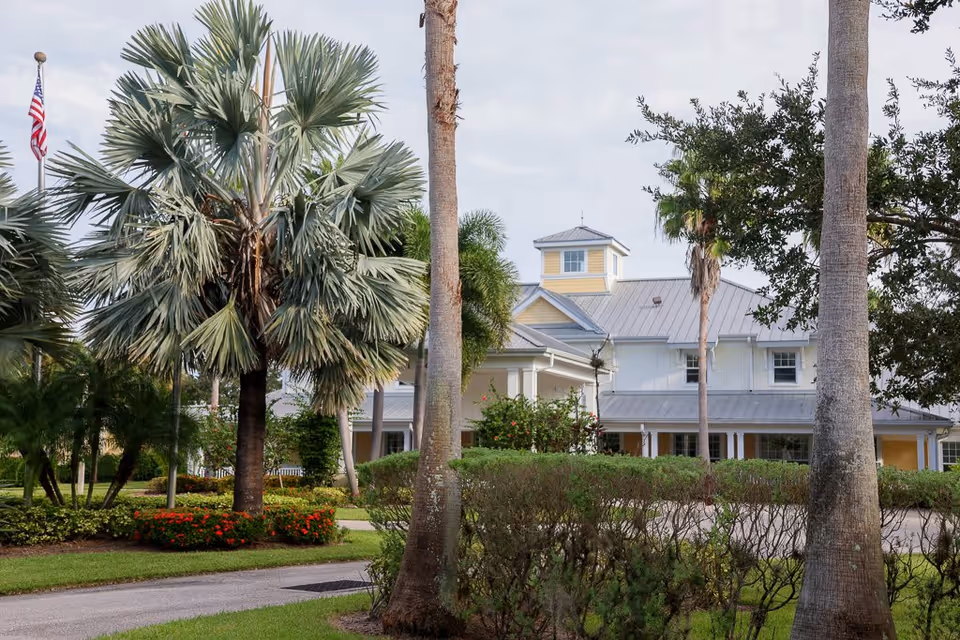 Exterior view of The Brennity at Tradition Senior Living building surrounded by palm trees, bushes, and a well-maintained garden with an American flag visible in the background.