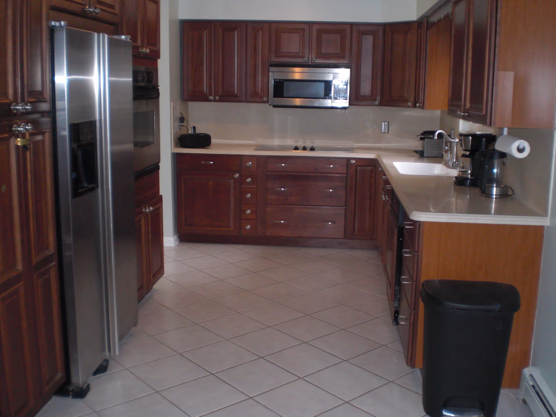 A tiled kitchen with dark wood cabinets, stainless steel refrigerator and microwave, countertops with sink and small appliances, and a trash bin.