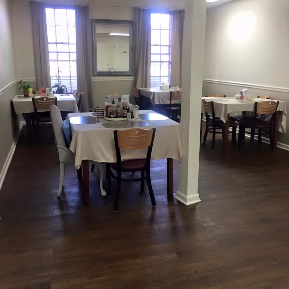 A dining room with several tables covered with white tablecloths, each surrounded by wooden chairs. The tables have condiments and napkin holders on them. There are two large windows with curtains and a mirror on the wall between them. The floor is wooden, and the walls are painted light beige with white trim.