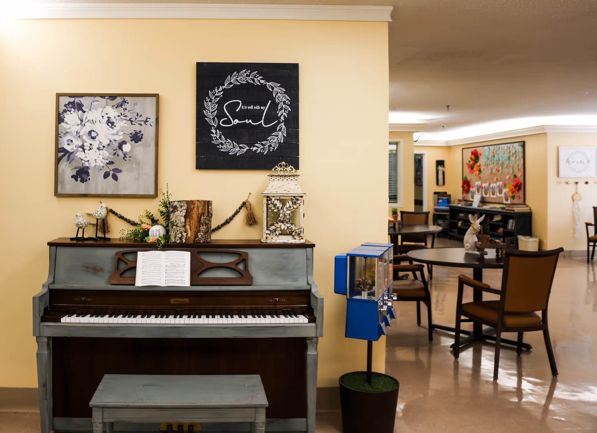 Interior view of a senior living facility common area featuring a vintage upright piano with an open music book, decorative items on top including a lantern and floral arrangement, and wall art above. In the background, there are round tables with chairs, a bulletin board with colorful decorations, and additional wall art. The space has a warm, inviting atmosphere with beige walls and polished floors.