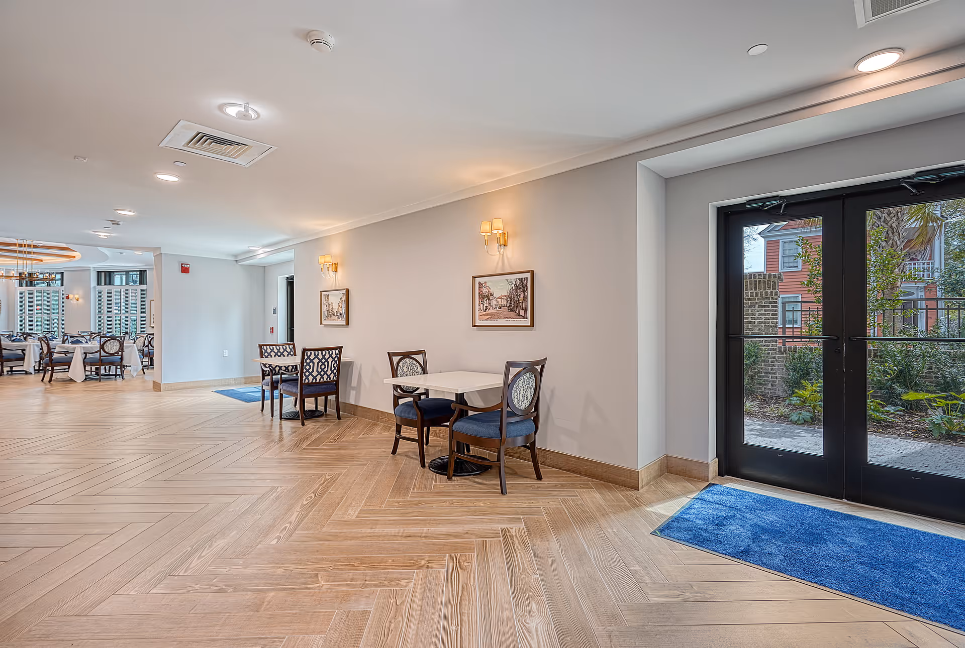 Interior view of a senior living facility with wooden herringbone patterned flooring, small tables with chairs along a wall, framed pictures, wall sconces, and large glass double doors leading outside to a garden area.