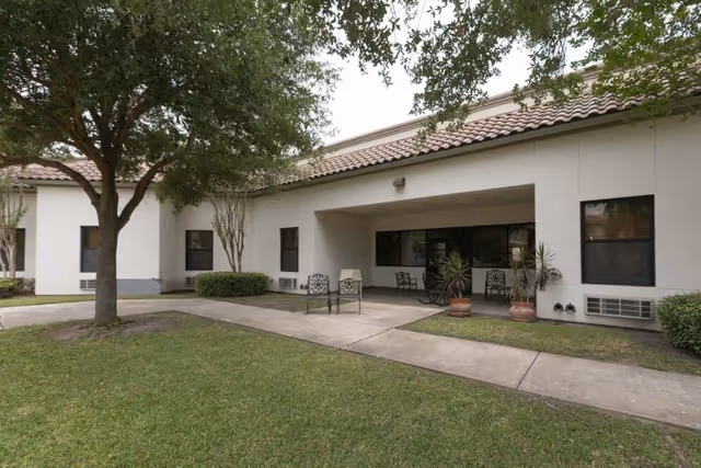 Single-story building front with a covered patio, metal chairs and potted plants beside a sidewalk and lawn under trees.
