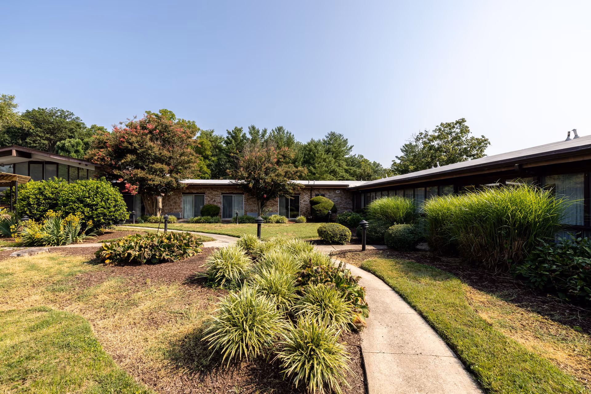 A landscaped outdoor courtyard area with a curved concrete pathway surrounded by green shrubs, bushes, and trees. The courtyard is enclosed by a single-story building with large windows and a flat roof under a clear blue sky.