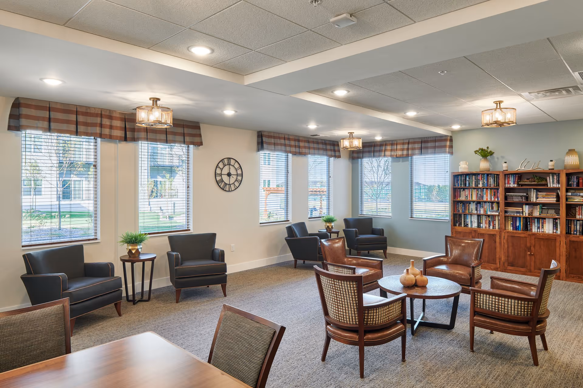 A well-lit community living room with multiple armchairs arranged around a circular wooden coffee table. The room features large windows with blinds and plaid valances, a wall clock, a wooden bookshelf filled with books and decorative items, and small side tables with potted plants. The ceiling has recessed lighting and decorative light fixtures.