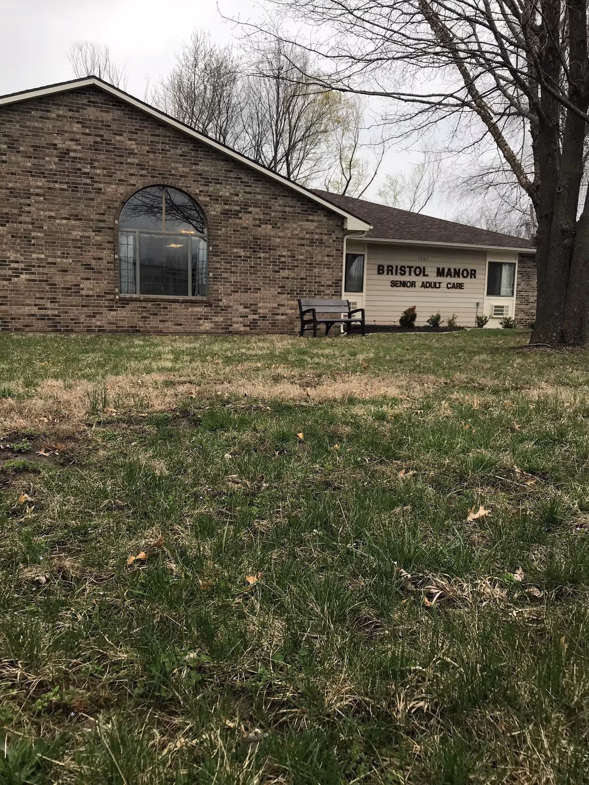 Exterior view of Bristol Manor Senior Adult Care building with a brick wall and large arched window, a bench on the lawn, and a tree on the right side under an overcast sky.