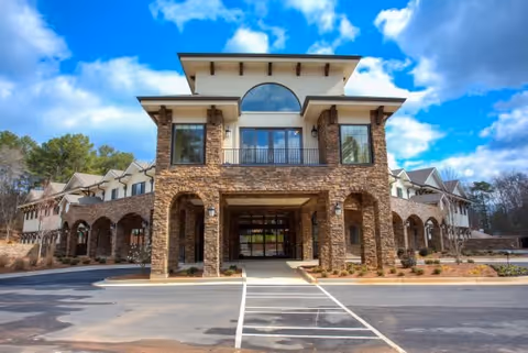 Front exterior view of Tapestry House Assisted Living at Alpharetta, featuring a large stone and stucco building with multiple windows, arches, and a covered entrance under a partly cloudy blue sky.