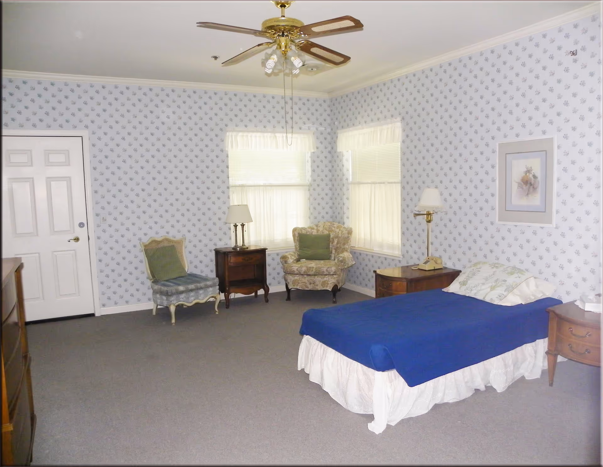 A senior retirement bedroom with a single bed covered in a blue blanket and white bed skirt. The room has floral wallpaper, two windows with white curtains, a ceiling fan with lights, two armchairs with cushions, two wooden nightstands with lamps, a rotary phone, and a framed picture on the wall.